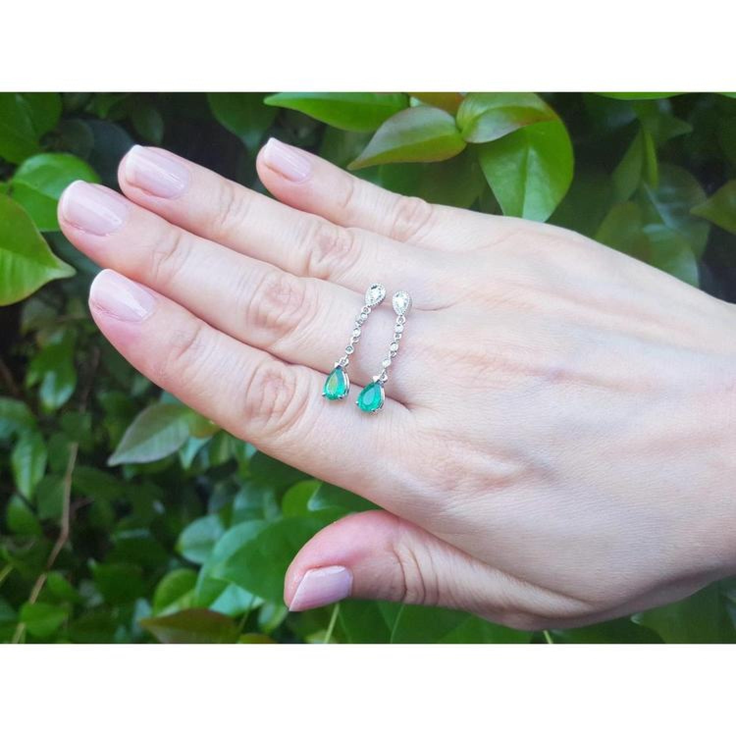 14K white gold pear-shaped emerald drop earrings with diamonds, held on a hand.