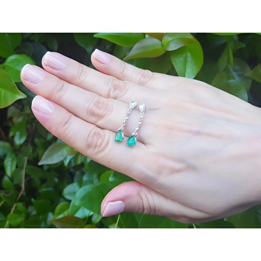 14K white gold pear-shaped emerald drop earrings with diamonds, held on a hand.