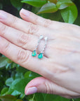 Hand displaying a pear shaped emerald and diamond earrings against a green leafy background