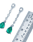 Pair of emerald teardrop earrings with diamond accents next to a ruler for scale on a white background