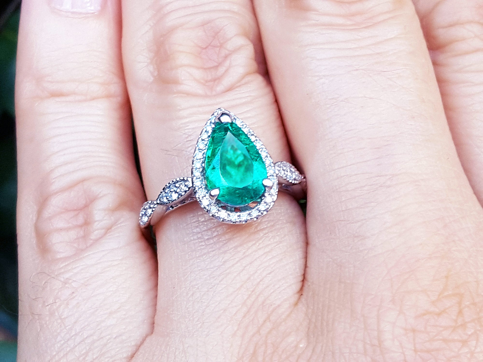 A teardrop emerald ring with a halo diamond on a finger.