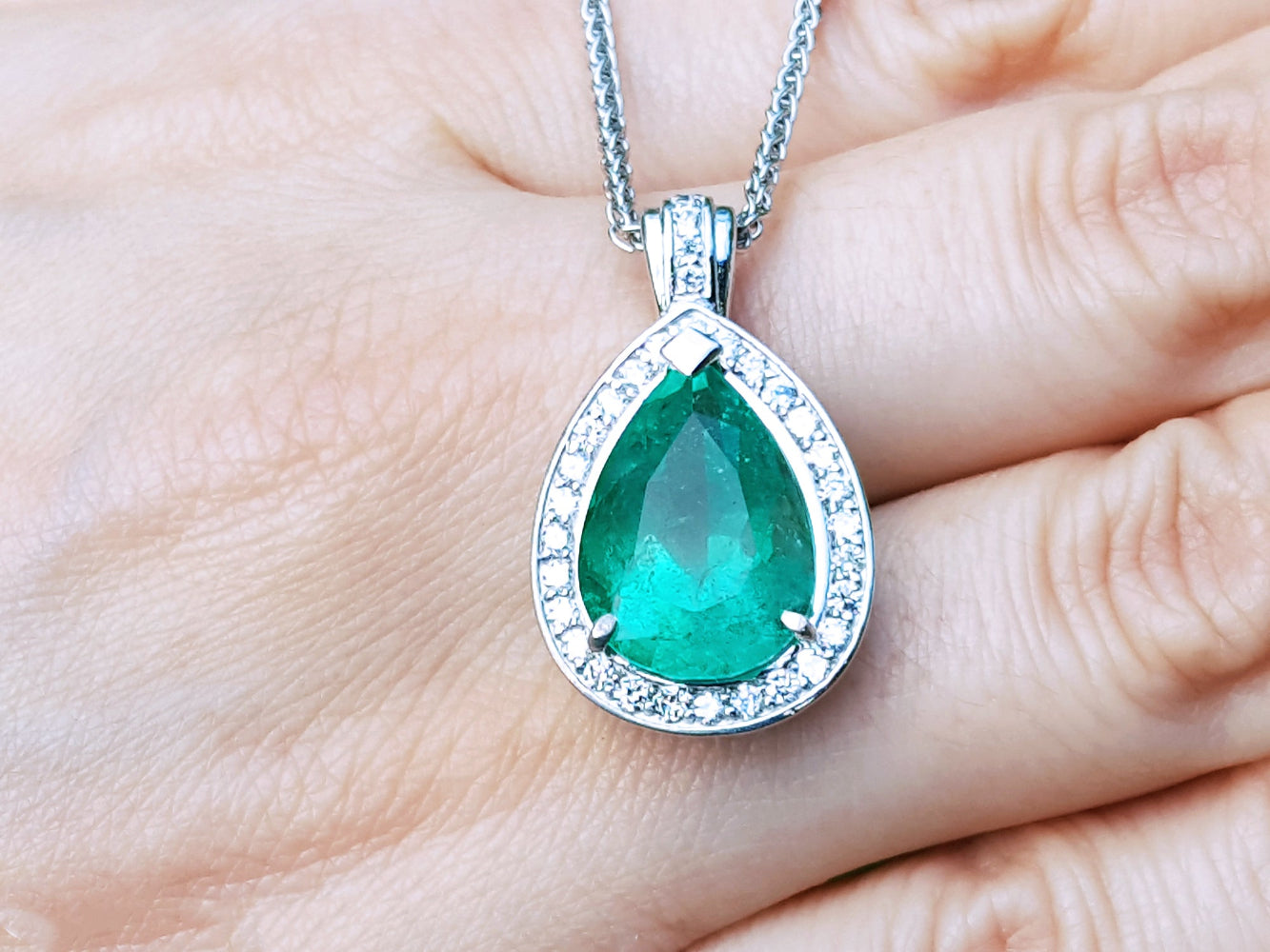 Green teardrop emerald gemstone pendant on a white gold chain held between fingers.