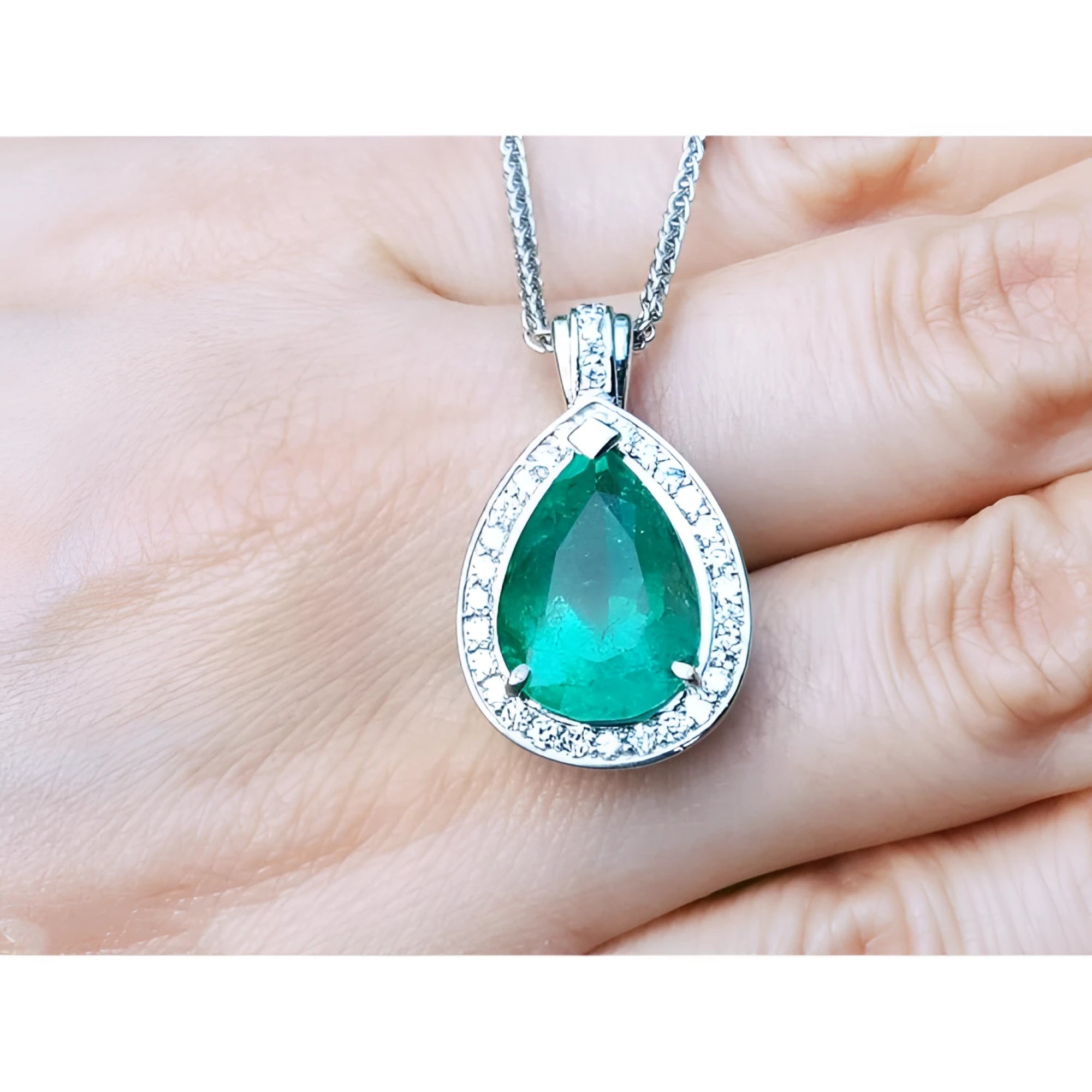 Pear-shaped emerald pendant with halo diamonds in 14K white gold, held by a hand.