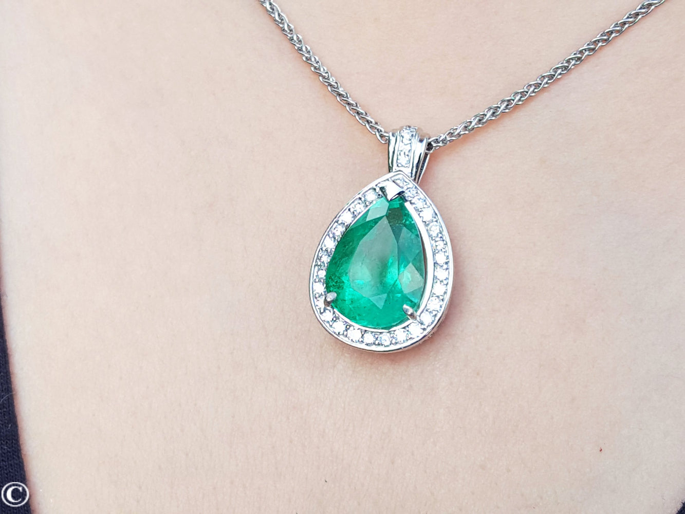 A white gold necklace with a natural emerald teardrop pendant on a beige skin background.