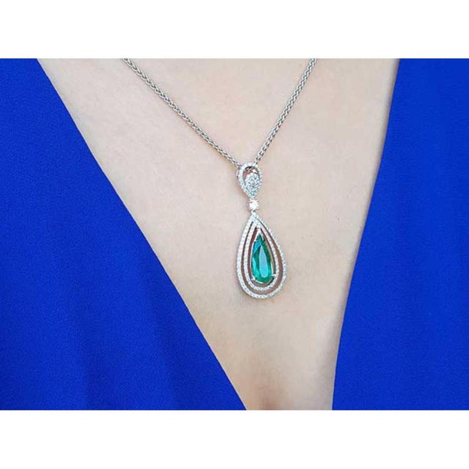 Teardrop emerald pendant with double diamond halo on an 18K white gold chain, worn on a blue dress.