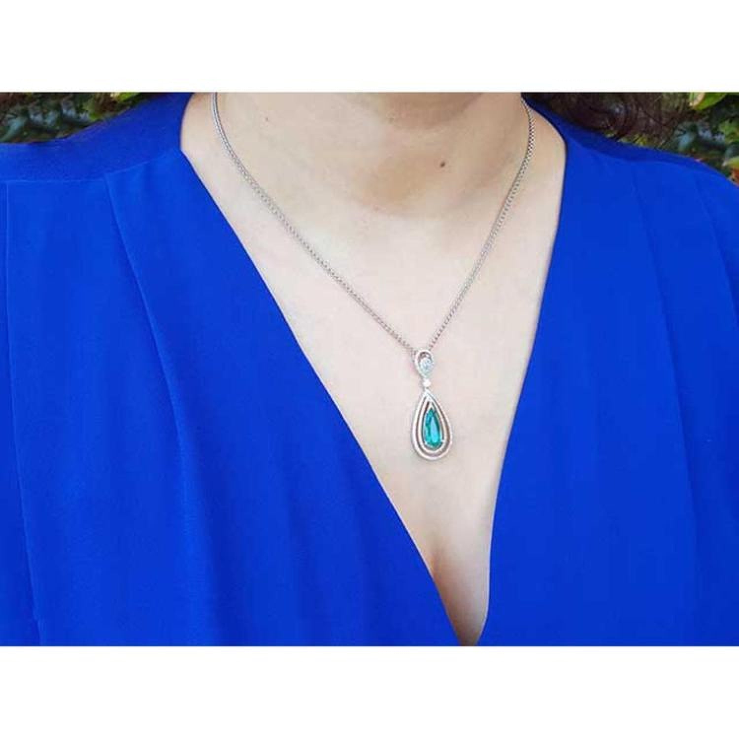Teardrop emerald pendant with double diamond halo on an 18K white gold chain, worn on a blue top.