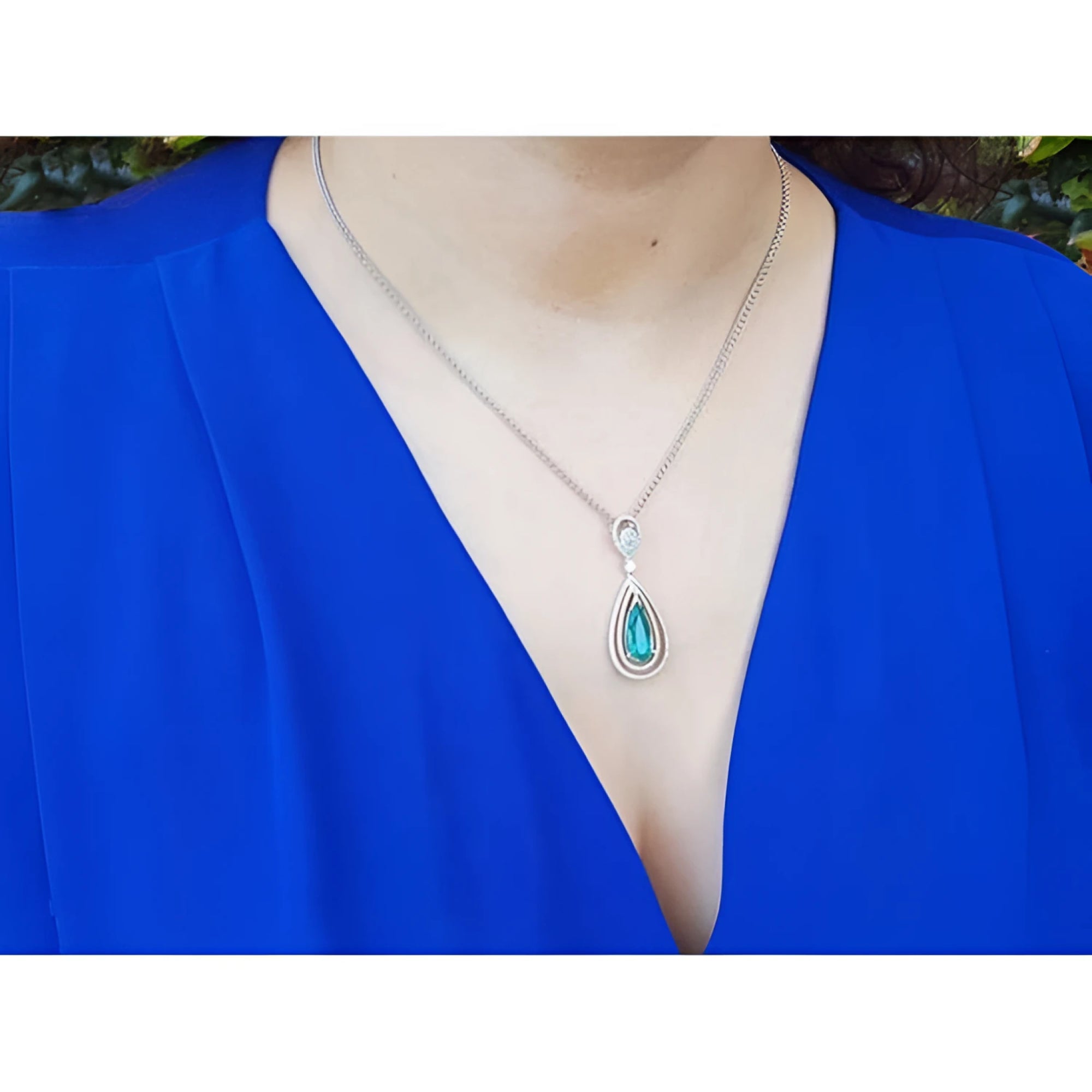 Teardrop emerald pendant with double diamond halo on an 18K white gold chain, worn on a blue top.