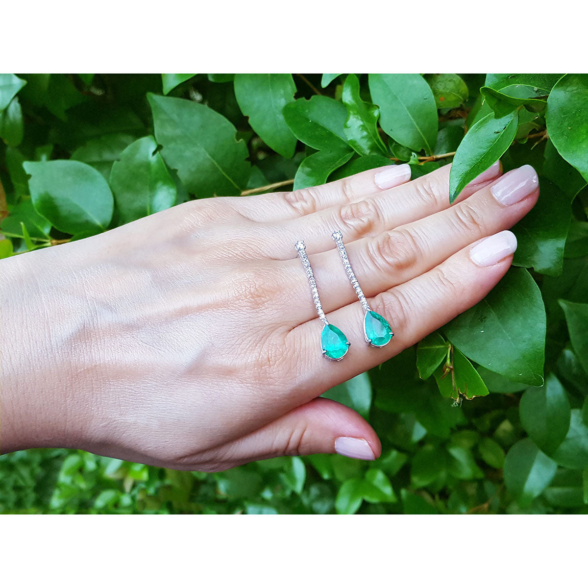 Emerald teardrop earrings on a hand with green leaves in the background