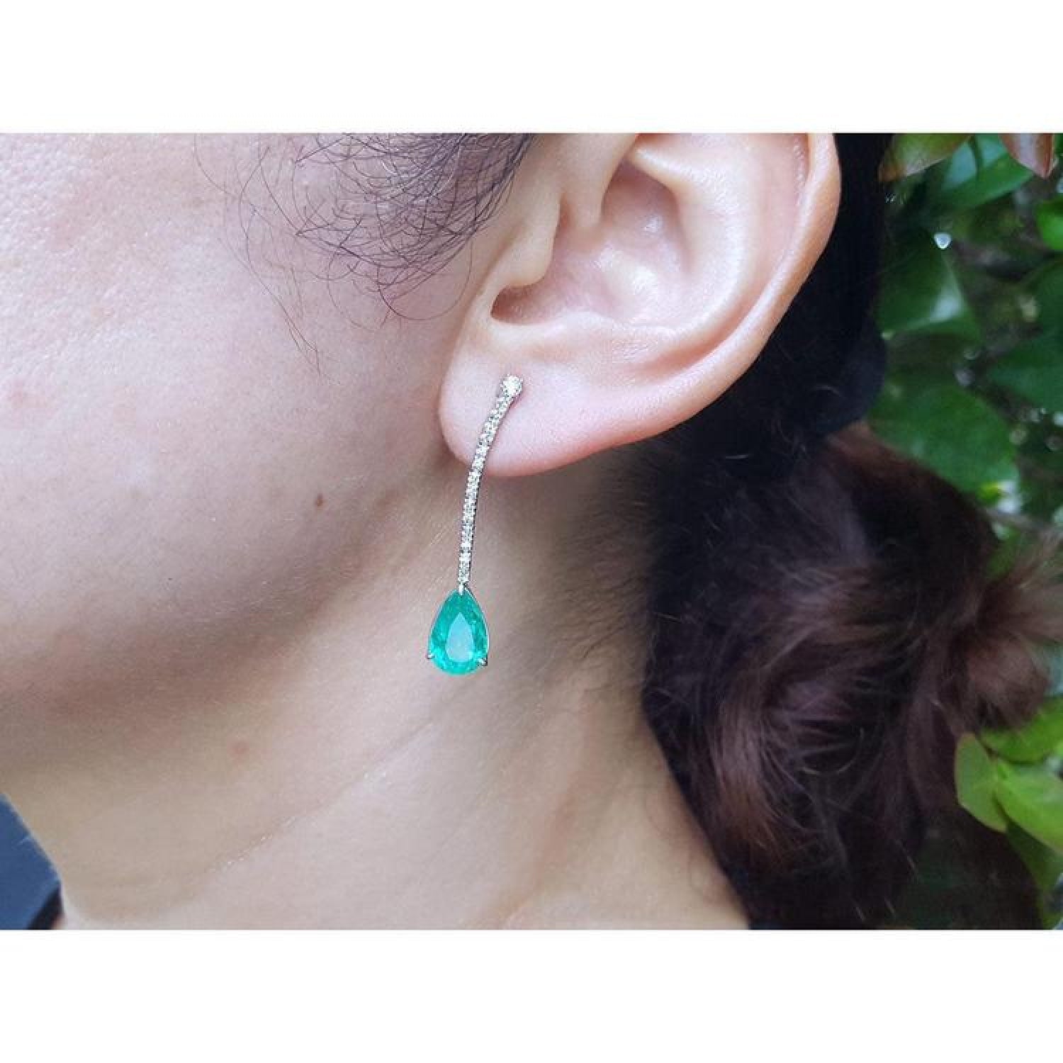 Pear shape 5.05 ct. emerald drop earrings in 18K white gold with diamond accents, worn on an ear.