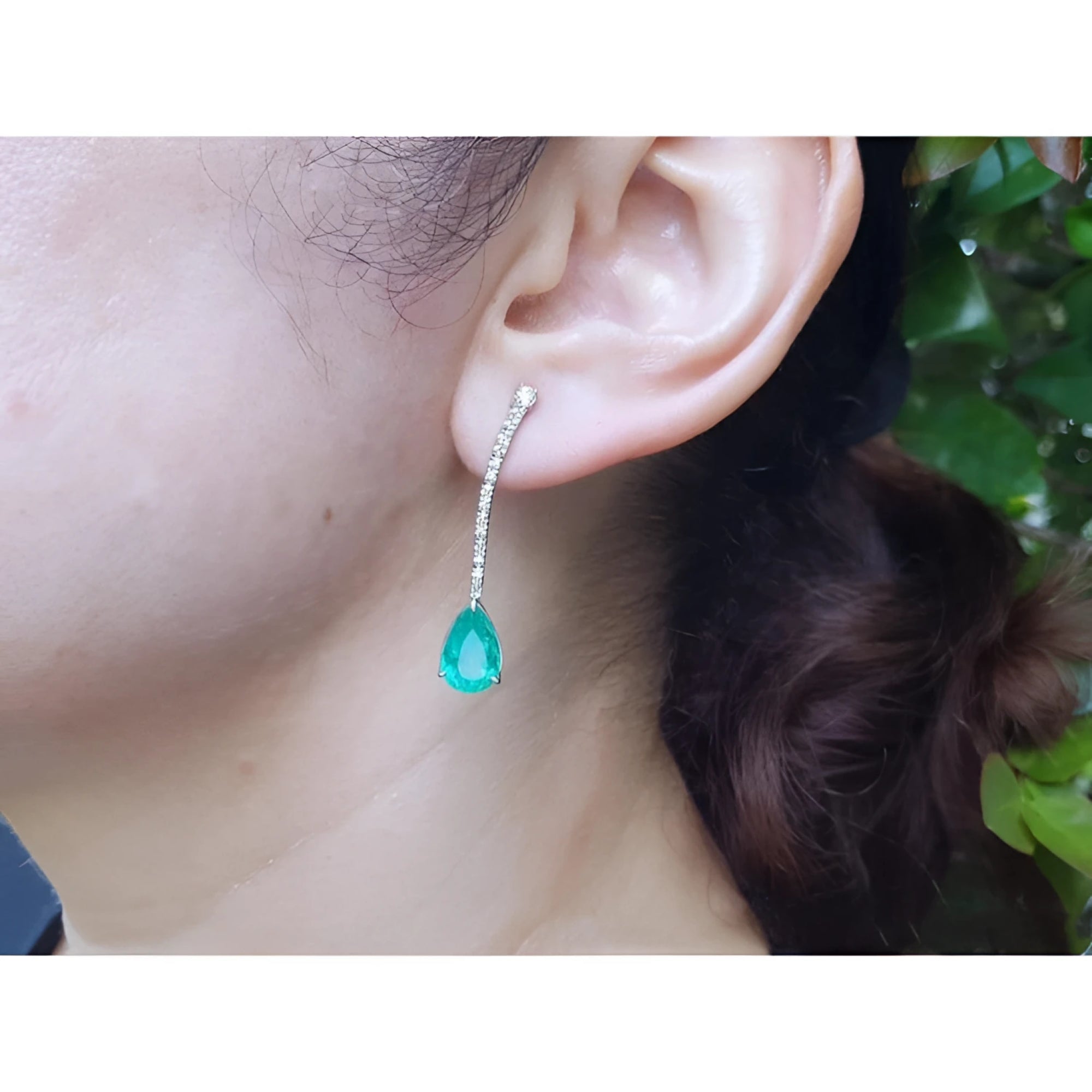 Pear shape 5.05 ct. emerald drop earrings in 18K white gold with diamond accents, worn on an ear.