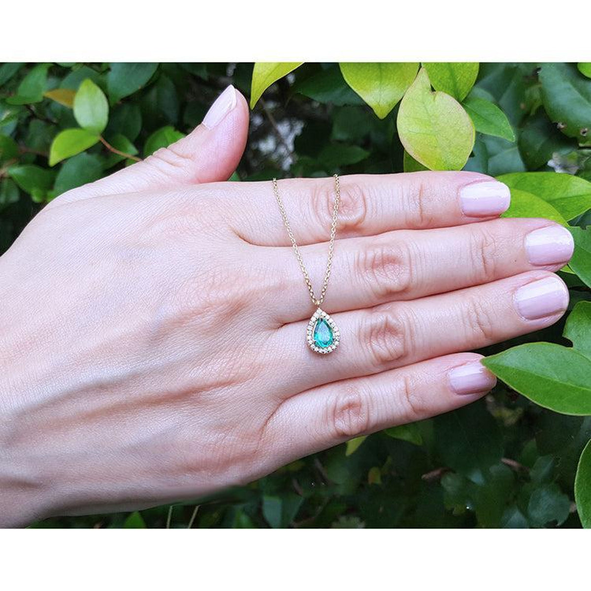 Pear-shaped emerald and diamond halo necklace in 14K yellow gold, held against green leaves.