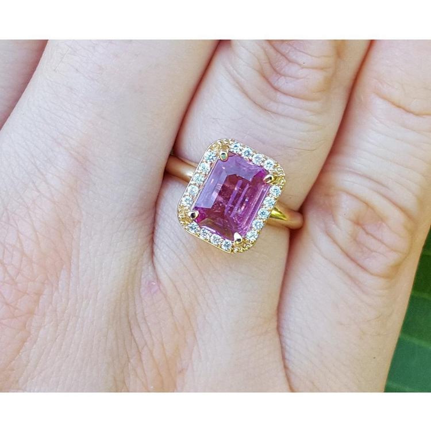Emerald-cut pink sapphire ring with diamond halo in 14K yellow gold on a hand.
