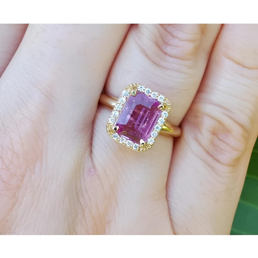 Emerald-cut pink sapphire ring with diamond halo in 14K yellow gold on a hand.