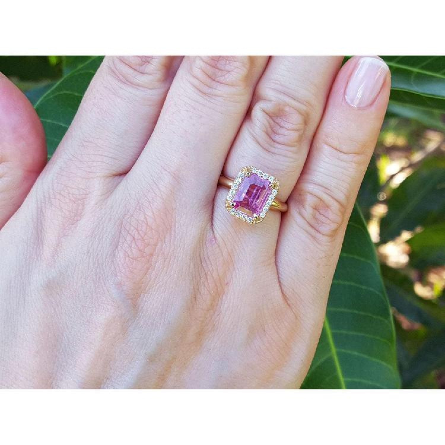 Emerald-cut pink sapphire ring with diamond halo in 14K yellow gold on a woman's hand.