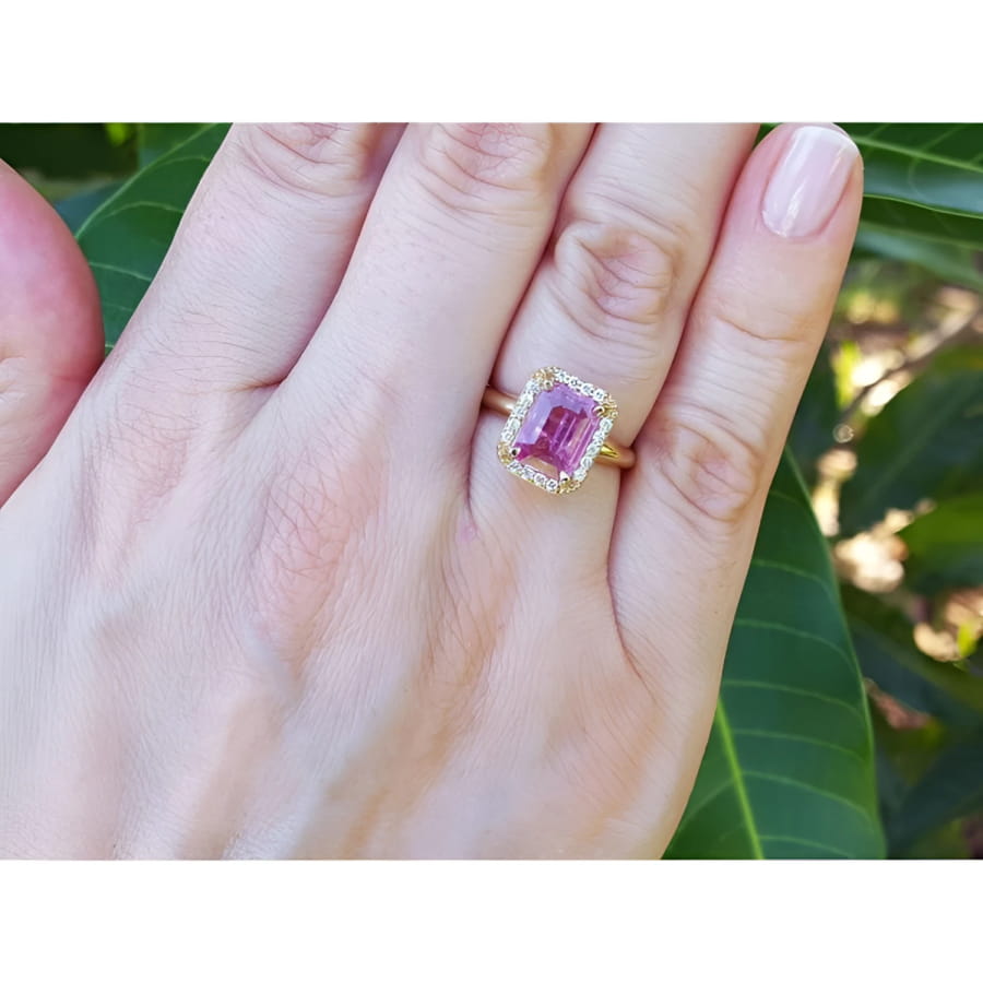 Emerald-cut pink sapphire ring with diamond halo in 14K yellow gold on a woman's hand.