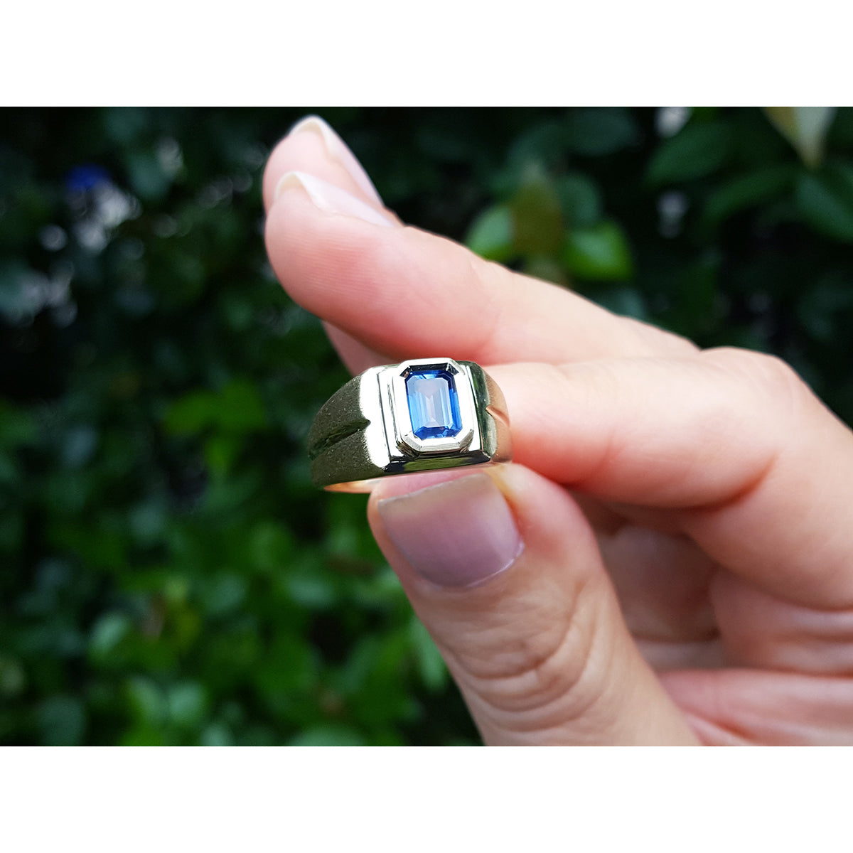 Between fingers holding a ring with a blue sapphire gemstone against a blurred green background