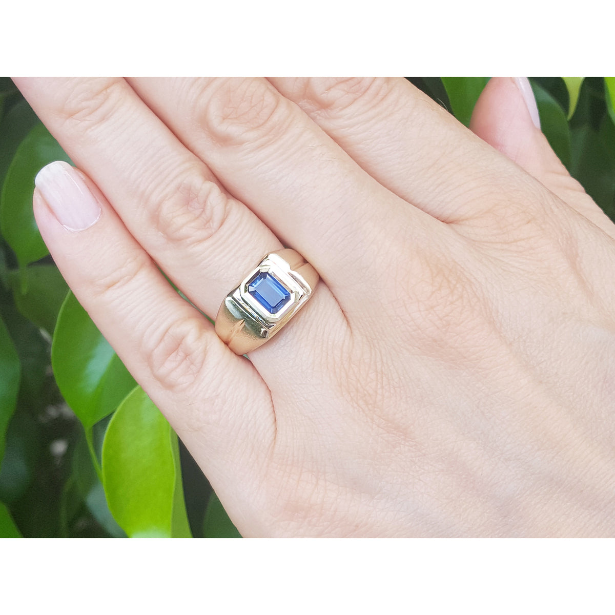 Gold ring with a blue sapphire gemstone on a hand against a green leafy background