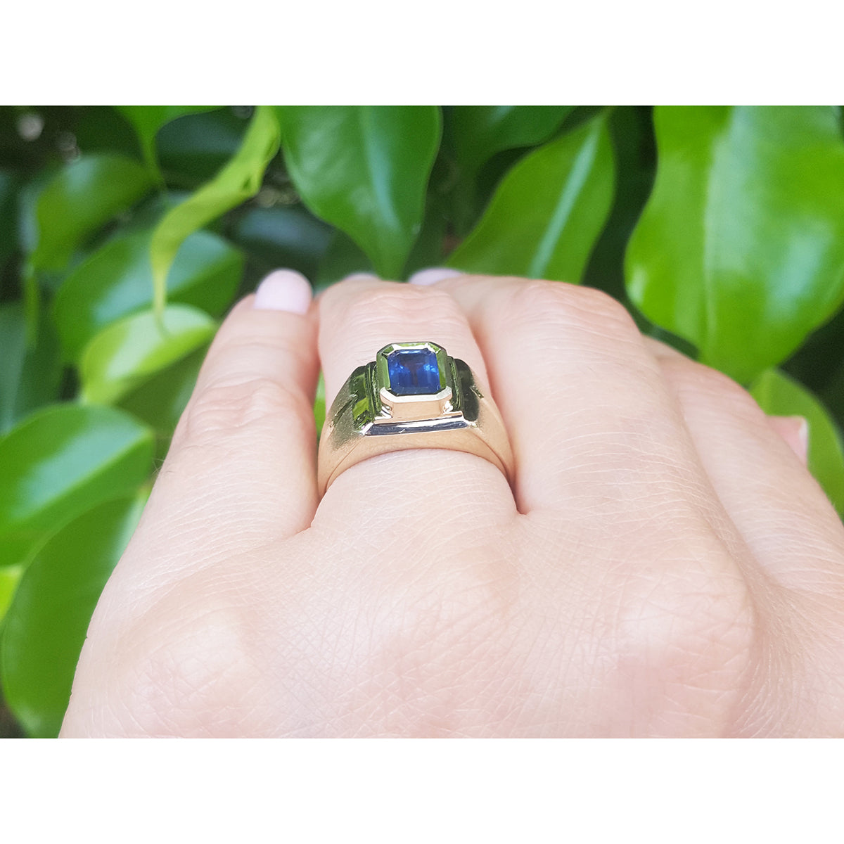 Gold solitaire ring with a blue sapphire gemstone on a hand against a green leafy background