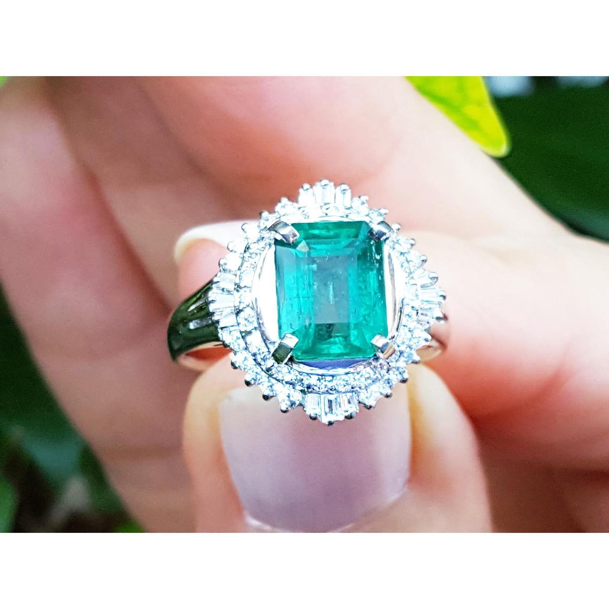 Emerald ring with diamond halo held in a hand against a blurred natural background.