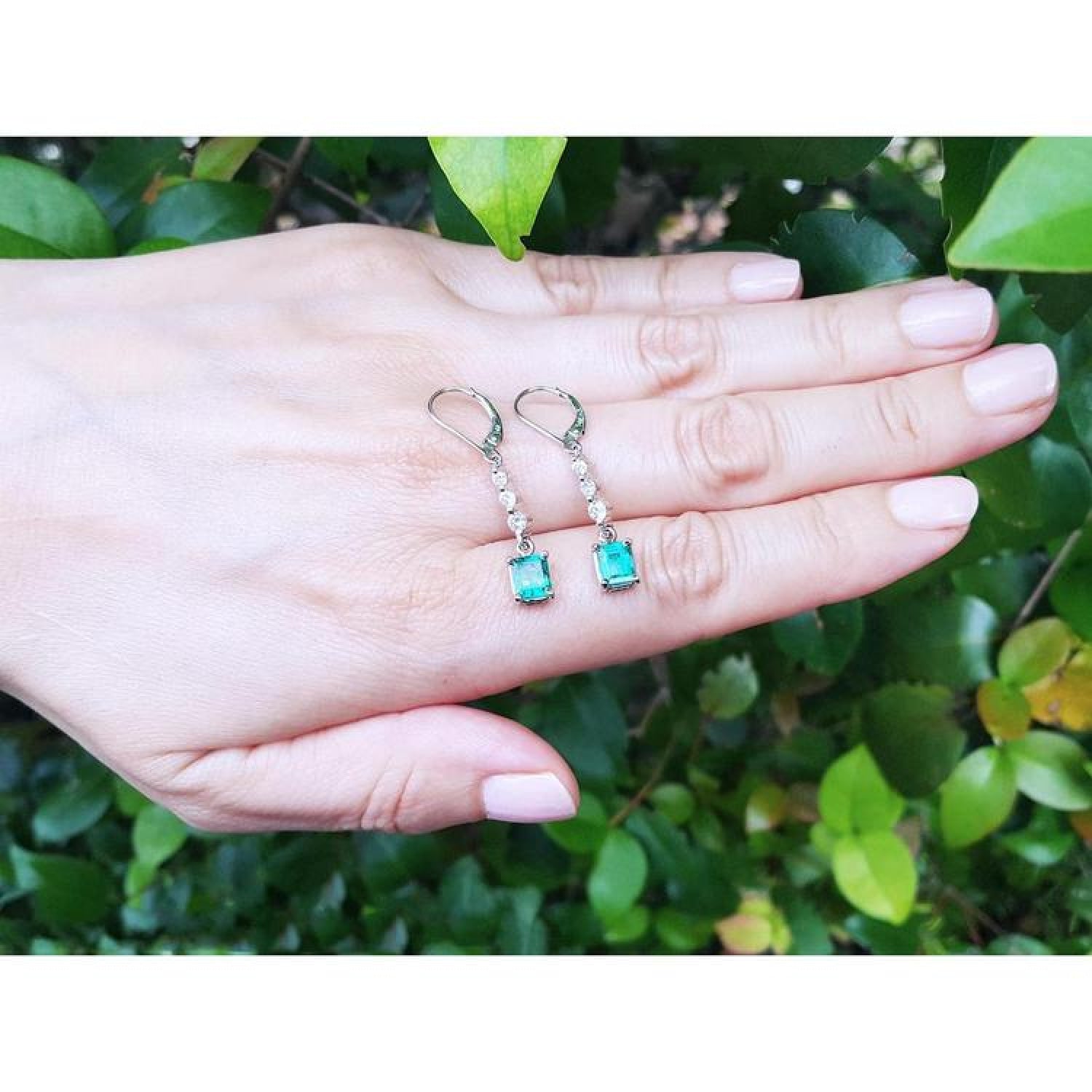 14K white gold dangle earrings with rectangular Colombian emeralds and clear stones on a hand.