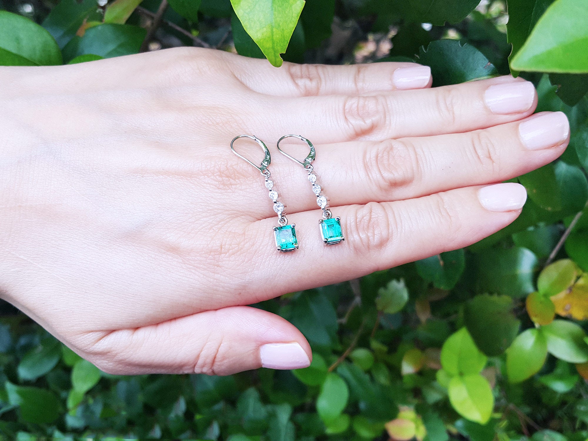 Pair of lever backs emerald dangle earrings held on a hand against a green leafy background