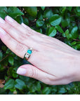 Hand wearing a gold mens ring with a real colombian emerald against a green leafy background.