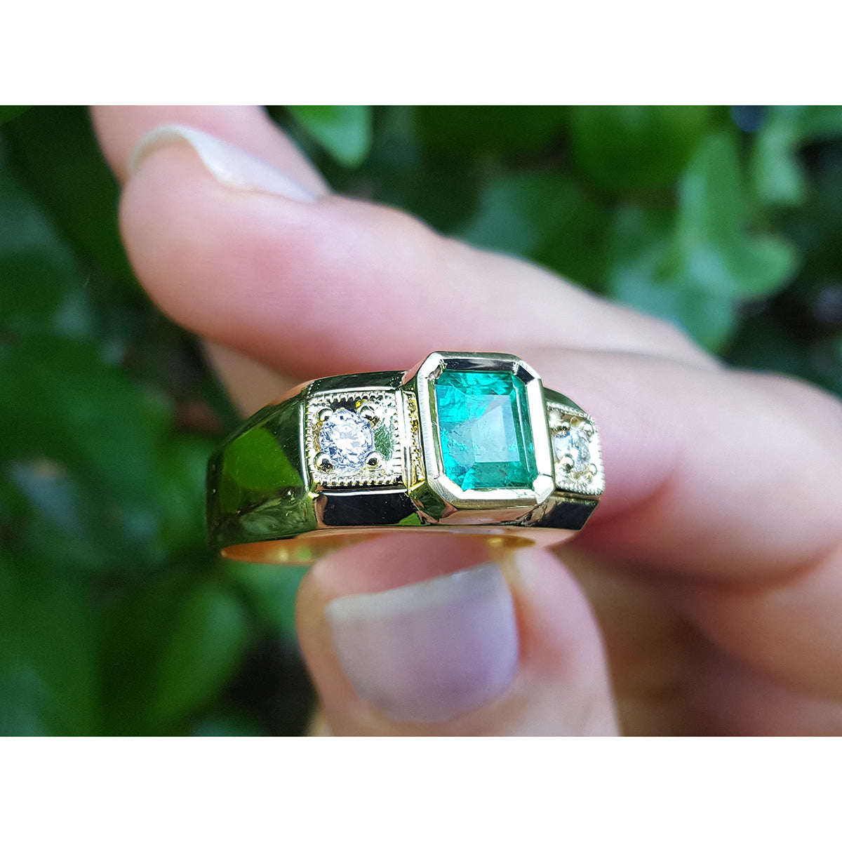 Gold mens ring with emerald and diamonds held between fingers against a green leafy background