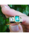 Gold mens ring with emerald and diamonds held between fingers against a green leafy background