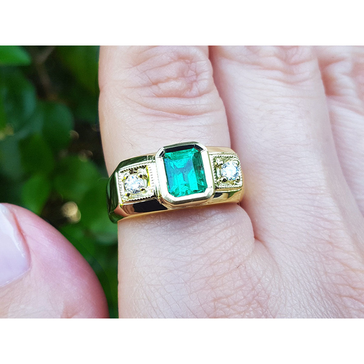 Gold ring with a square natural emerald on a finger against a blurred natural background.