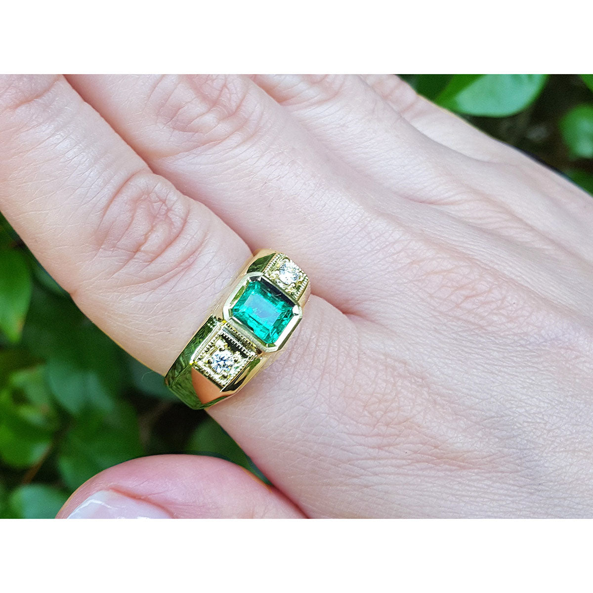 Gold ring for men with emerald and diamonds on a finger against a natural background.