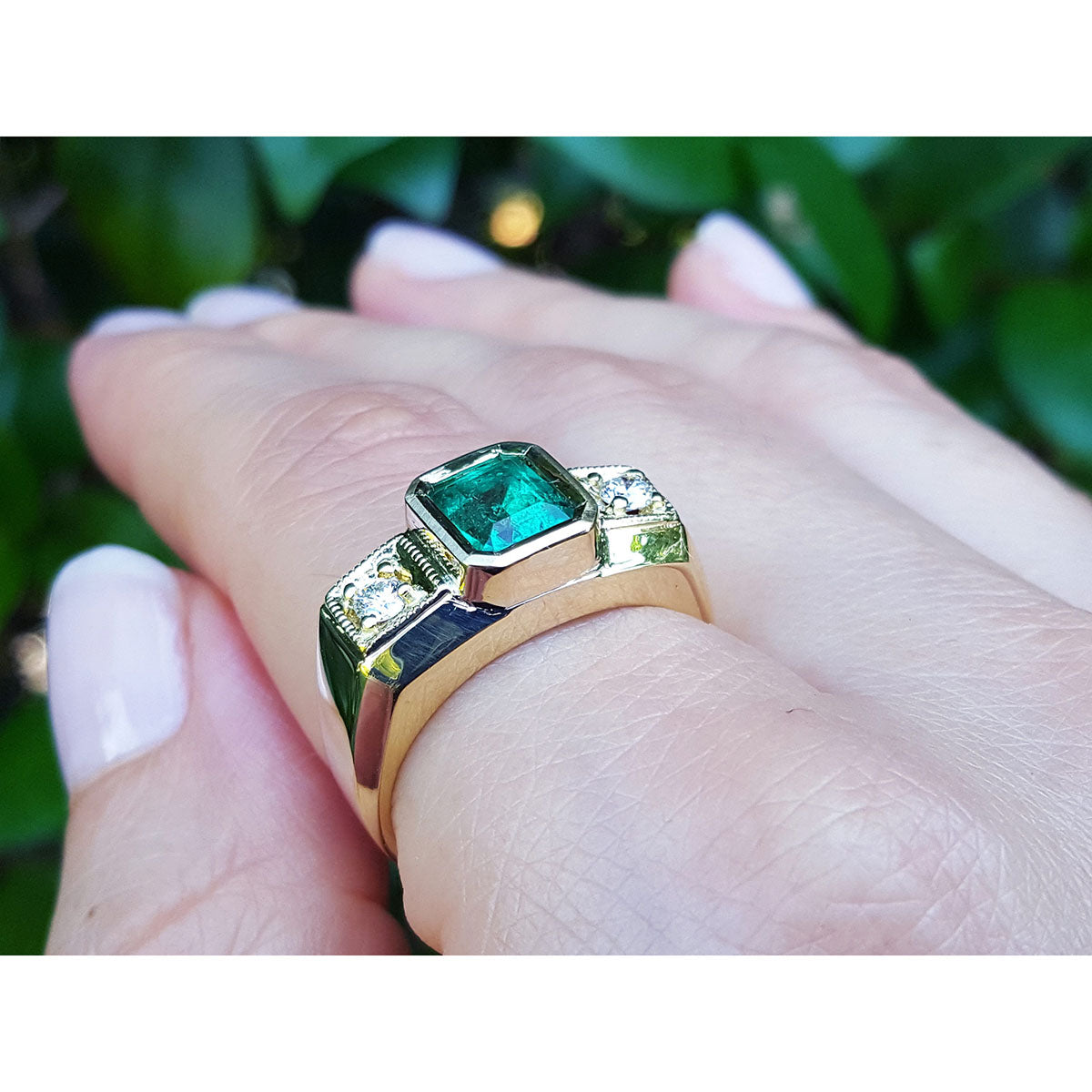 A men&#39;s gold ring with emerald and diamond on a hand against a blurred green background.