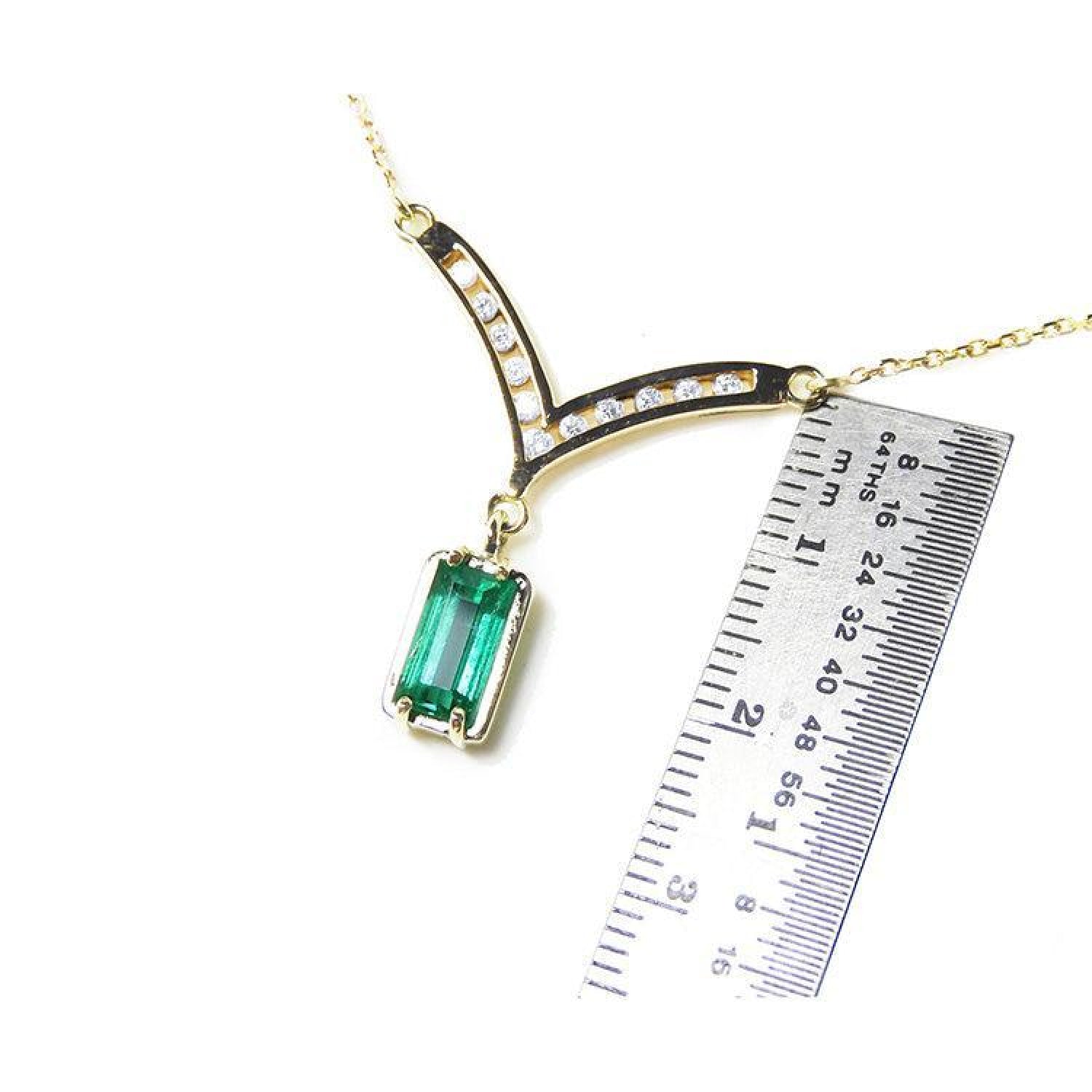 14K yellow gold necklace with a baguette-cut Colombian emerald pendant dangling from a diamond-accented curved V, shown next to a ruler for scale.