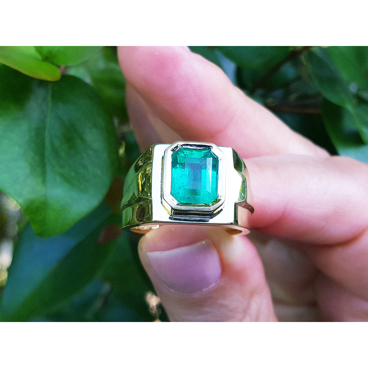 Gold solitaire ring with a rectangular emerald cut held between fingers against a green leafy background.