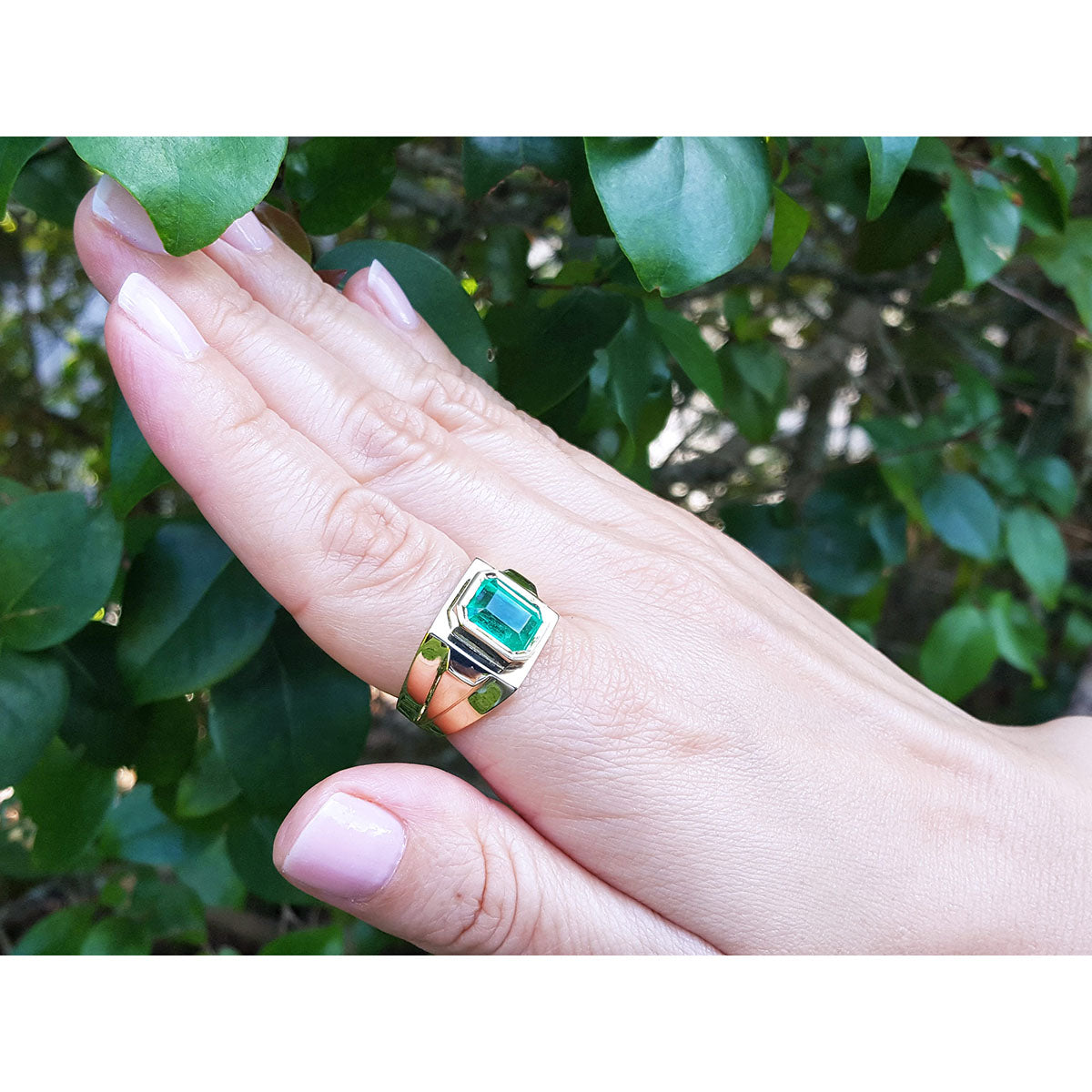 Hand wearing a ring with a genuine emerald gemstone against a leafy background.