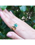 Hand wearing a ring with a genuine emerald gemstone against a leafy background.