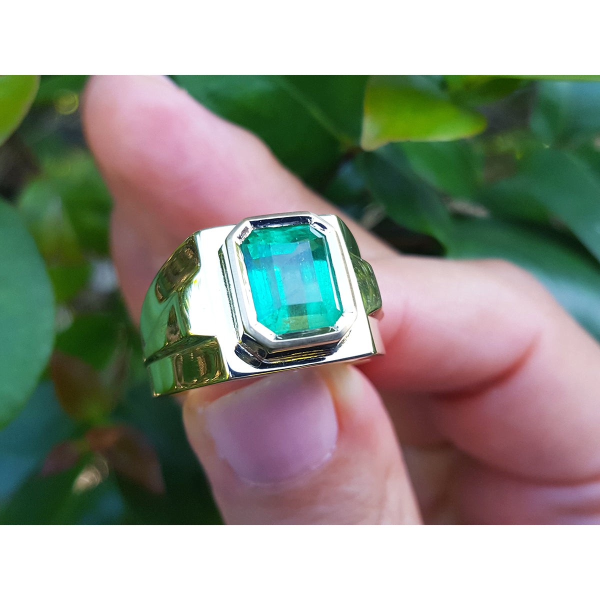 Gold solitaire ring with a bezel set emerald held by a hand against a blurred green leafy background.