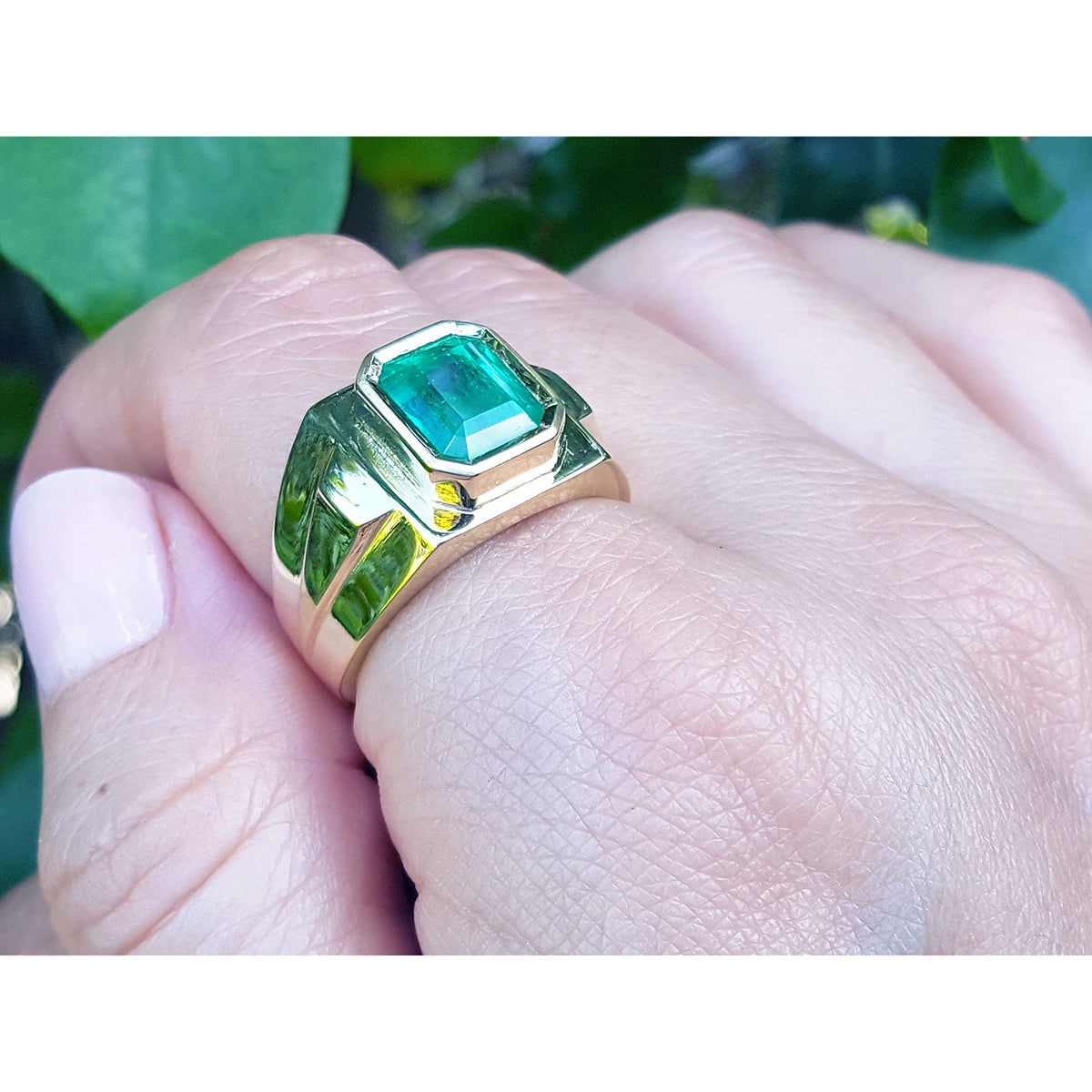A 18k yellow gold ring with a Muzo colombian emerald on a hand against a natural background.