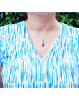 Person wearing a blue and white patterned shirt with a watermelon tourmaline necklace against a green leafy background.