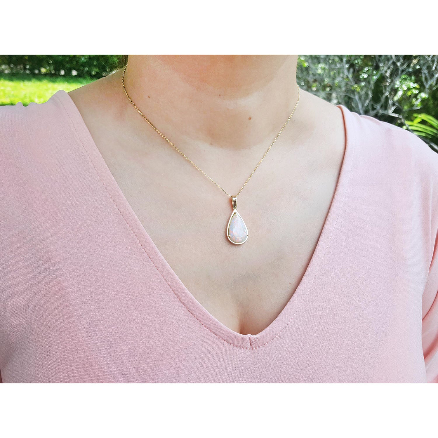 A woman wearing a gold necklace with a teardrop-shaped opal pendant against a blurred green outdoor background.