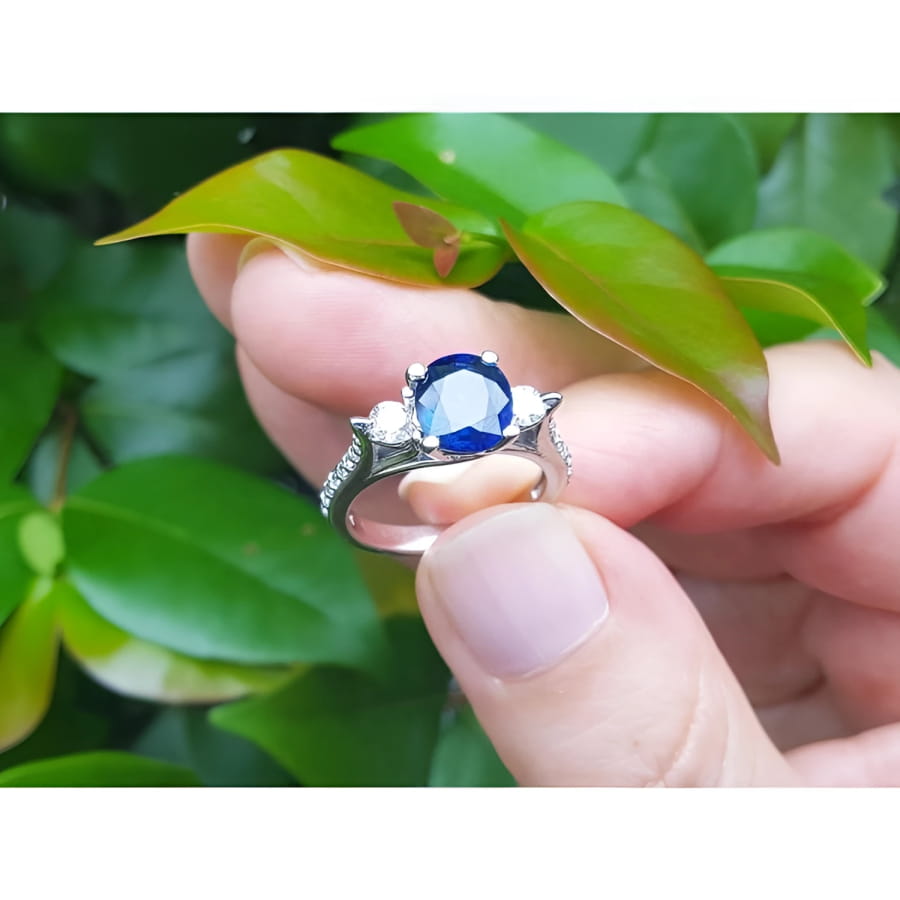 14K white gold ring with a deep blue oval sapphire and two side diamonds, held against green leaves.
