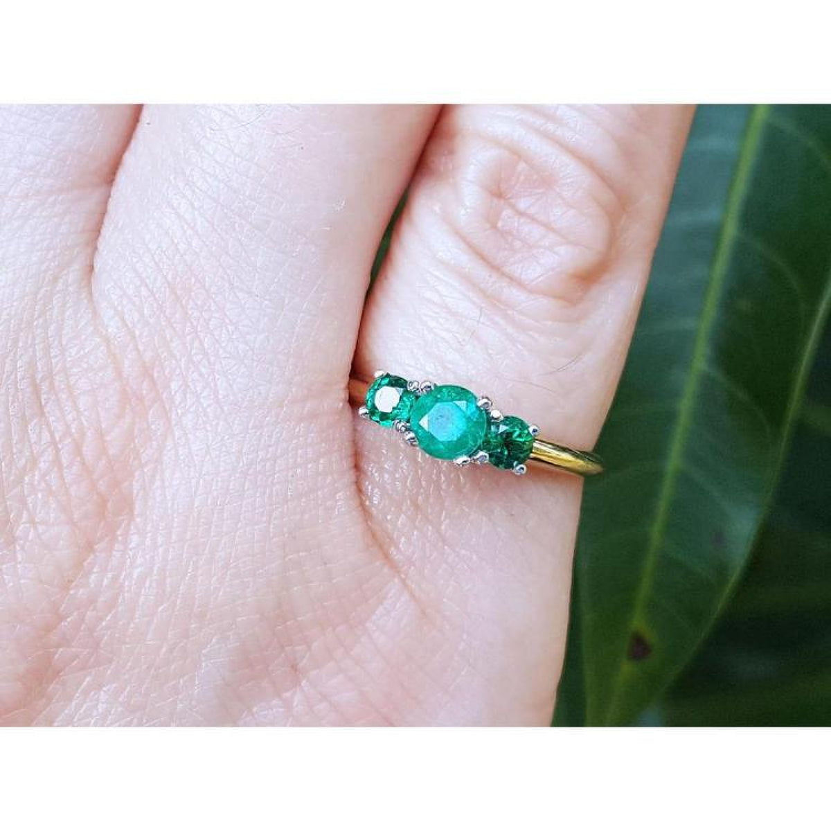Round cut three-stone emerald ring with prong-set stones in white and yellow gold on a finger.