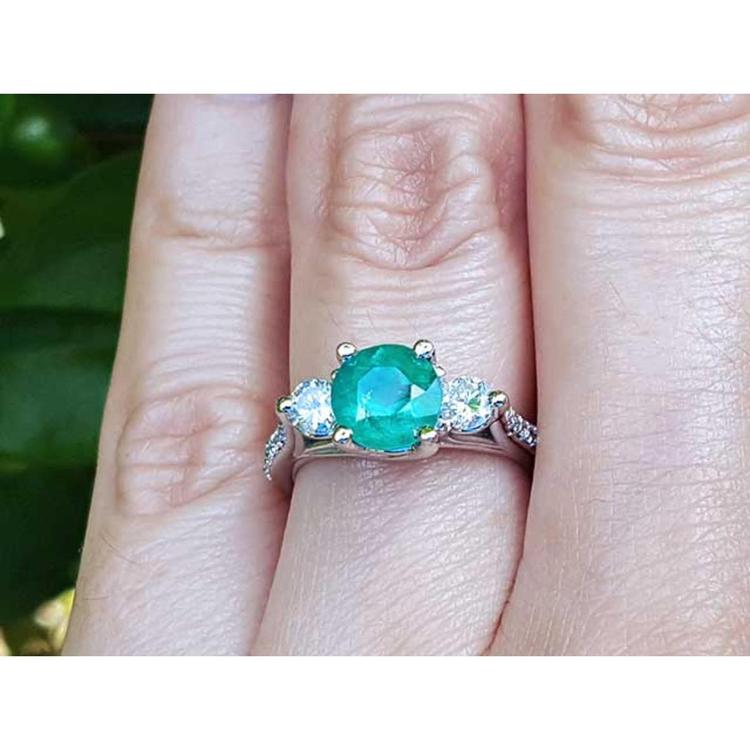 Emerald ring with side natural diamonds on a finger with a blurred green background.