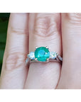 Emerald ring with side natural diamonds on a finger with a blurred green background.