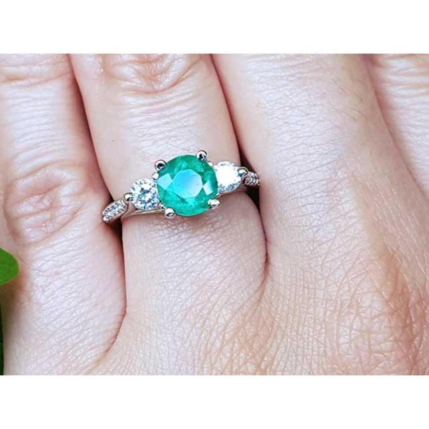 Round cut emerald and two side diamonds in a 14K white gold three-stone ring on a finger.