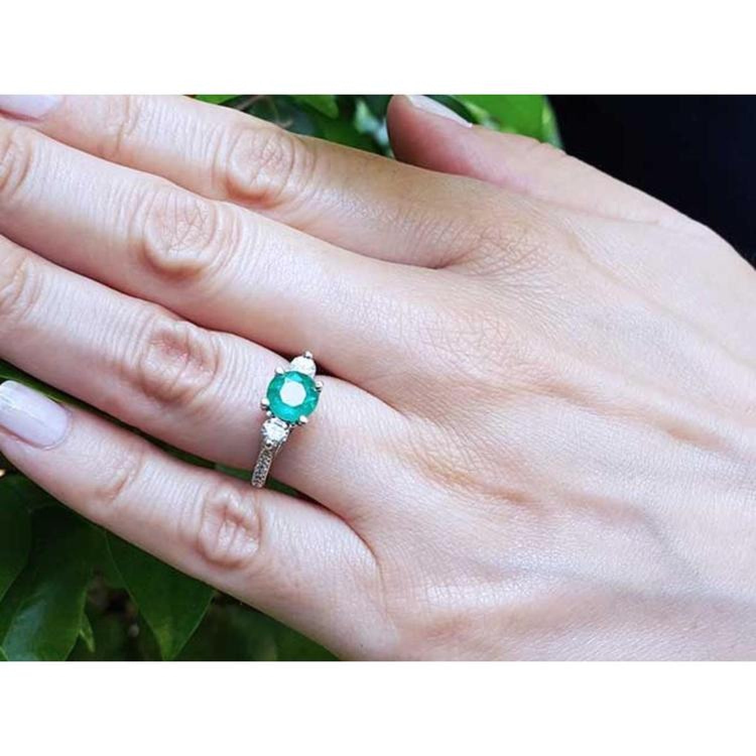 Round cut emerald ring with side diamonds in 14K white gold on a hand.