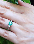 Hand wearing a ring with an emerald stone against a green leafy background.