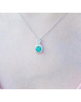Round Colombian emerald and diamond halo pendant in 18K white gold, worn on a woman's neck.