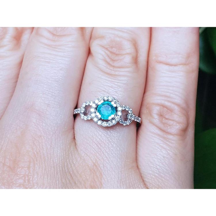 A round cut natural emerald surrounded by a halo diamond in 14k white gold on a finger.