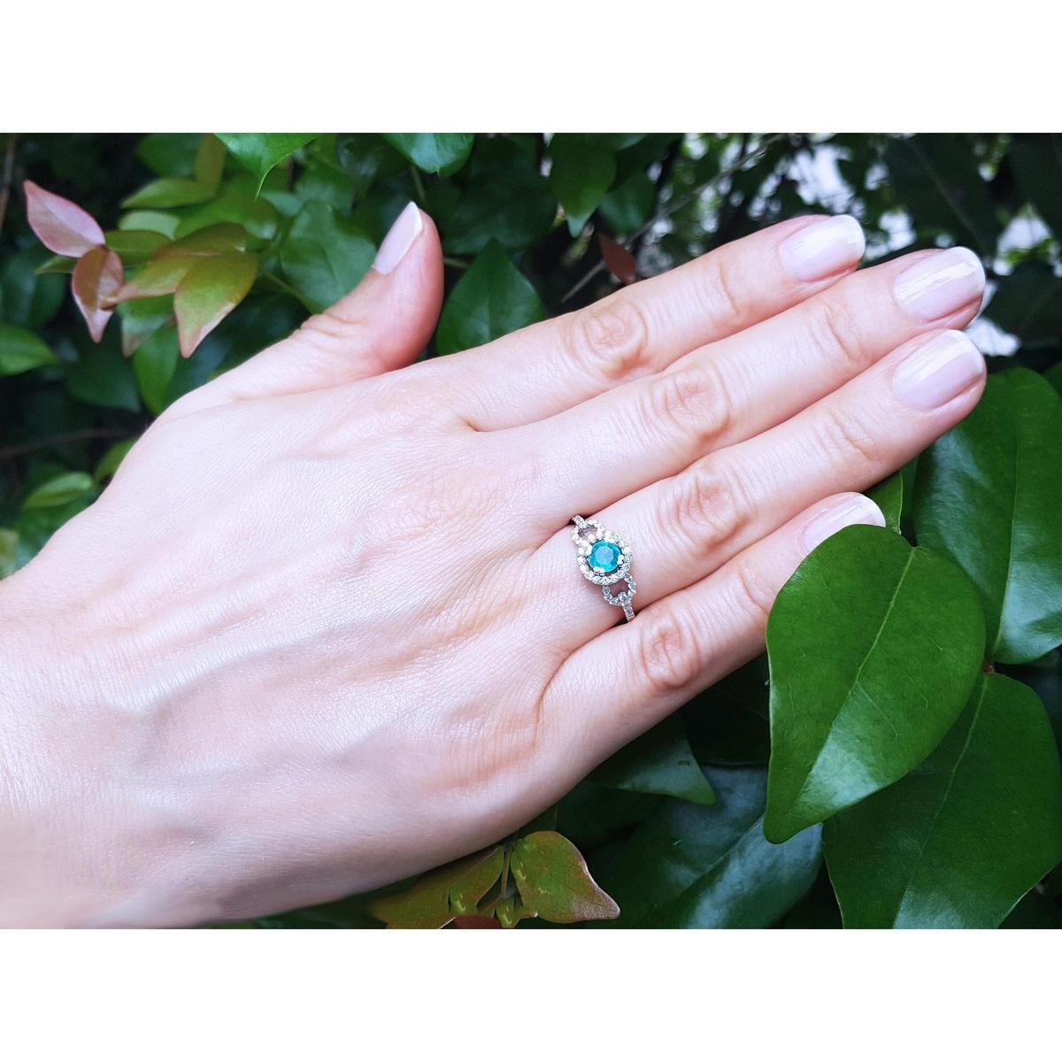 Hand wearing a ring with a round cut emerald against a leafy background.