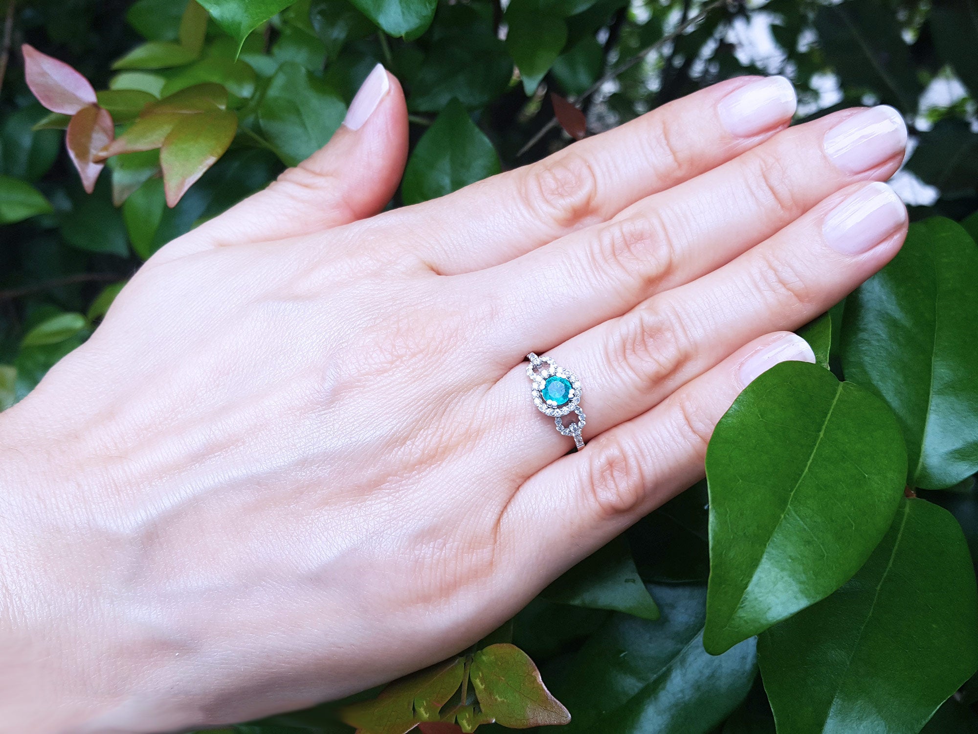 Hand wearing a ring with a round cut emerald against a leafy background.